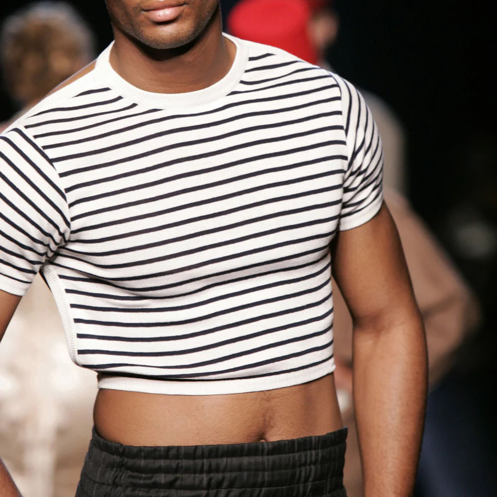 Model mit gestreiftem, bauchfreiem T-Shirt in Schwarz-Weiß auf einem Laufsteg, Fokus auf Oberkörper.