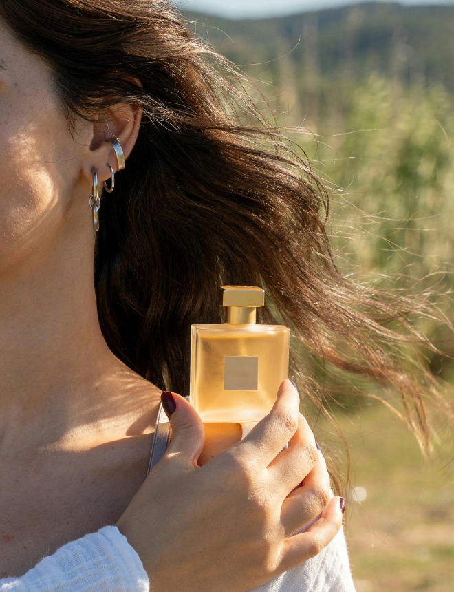Frau mit im Wind wehenden Haaren hält goldenen Parfümflakon vor herbstlicher Landschaft