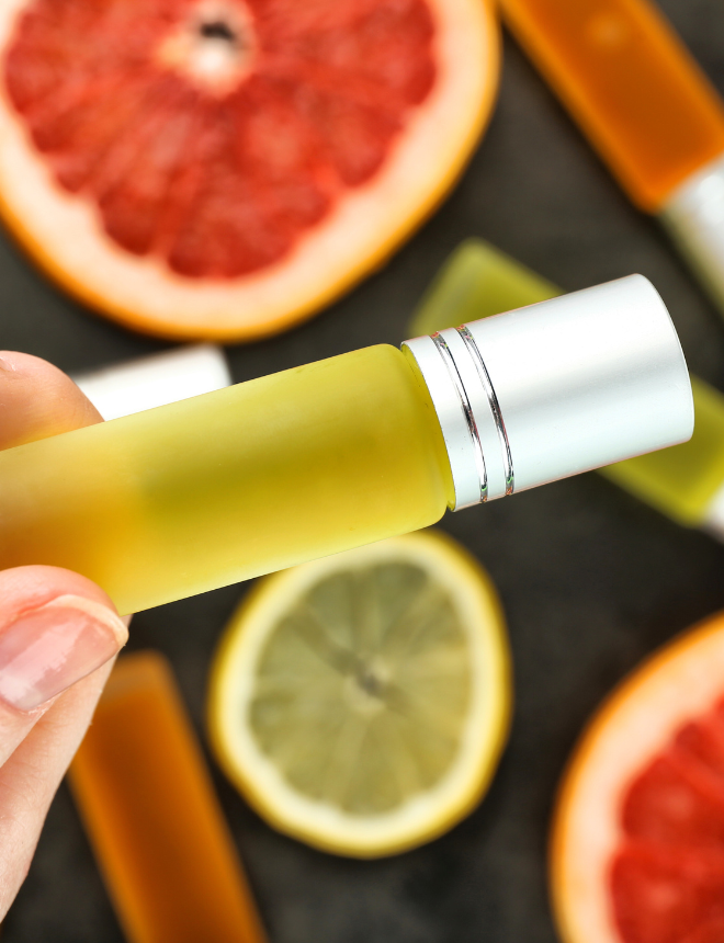 Hand hält gelbes Duftöl-Roller-Fläschchen vor aufgeschnittener Grapefruit und Zitrone, Sommerdüfte fruchtig