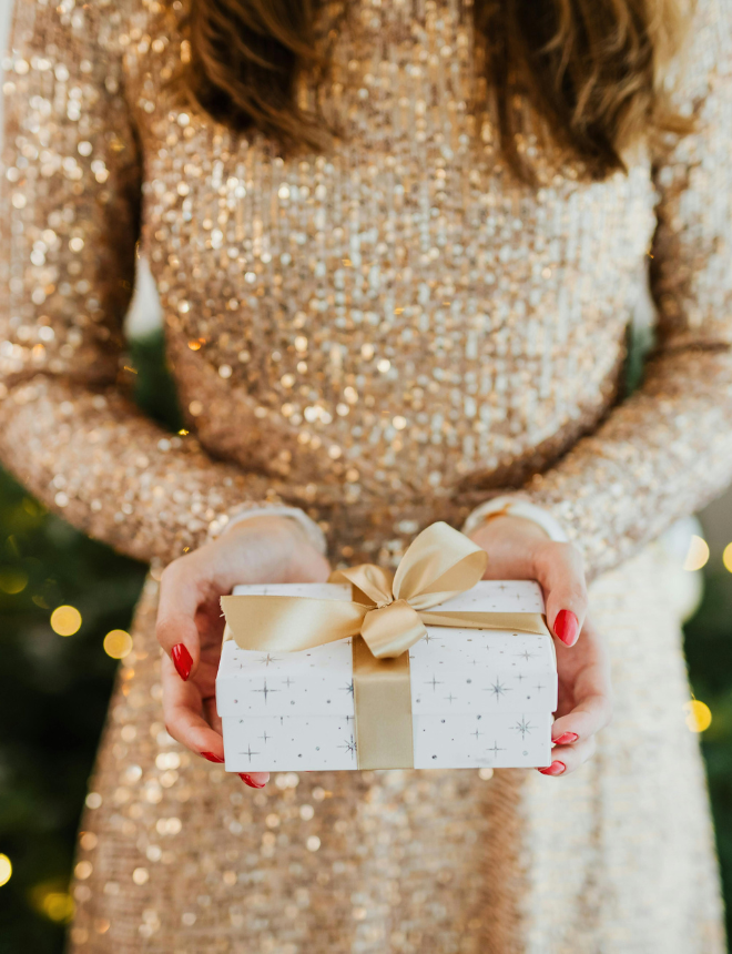 Frau im goldenen Glitzerkleid hält ein weiß verpacktes Geschenk mit goldener Schleife, Artikelbild Geschenkideen zu Weihnachten