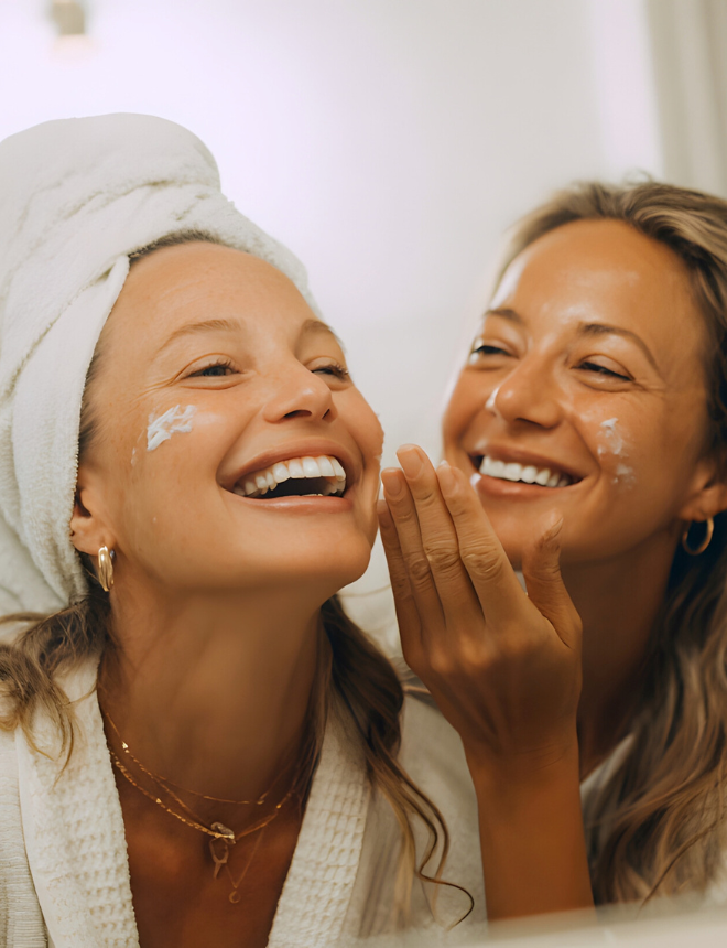 Zwei lachende Frauen tragen sich gegenseitig Gesichtscreme auf, Artikelbild Geschenkideen zu Weihnachten