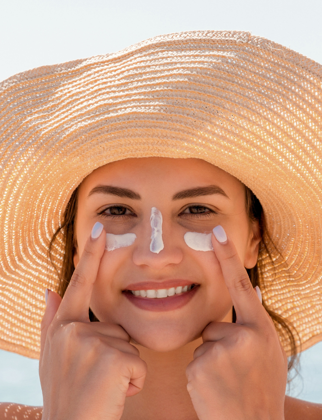 Lächelnde Frau mit Sonnenhut trägt Sonnencreme im Gesicht auf, Artikelbild Sonnenschutz