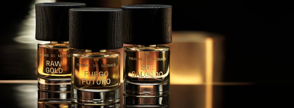 Drei Parfumflakons von Thomas de Monaco Parfums mit dunklen Holzverschlüssen, beschriftet Raw Gold, Fuego Futuro und Sol Salgado, auf glänzender Oberfläche