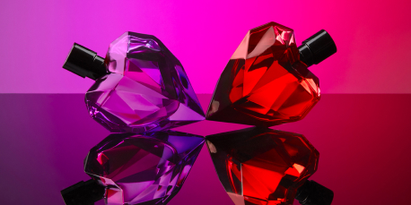 Zwei herzförmige Parfumflakons liegen sich gegenüber: einer in Violett, der andere in Rot. Beide haben eine facettierte, diamantähnliche Oberfläche und schwarzen Verschluss. Der Hintergrund verläuft in kräftigen Pink- und Violetttönen, während sich die Flakons spiegelnd auf einer glänzenden Oberfläche abzeichnen.