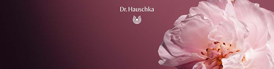 Werbebild von Dr. Hauschka: Nahaufnahme einer zartrosa Blüte mit weichen, gefalteten Blütenblättern vor einem roséfarbenen Hintergrund. Links oben steht das Dr. Hauschka-Logo in Weiß – das Motiv vermittelt natürliche Schönheit und Sanftheit.