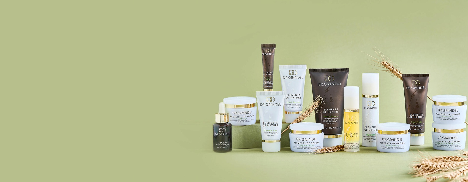 Dr. Grandel Elements of Nature Serie mit zahlreichen Pflegeprodukten in weißen und braunen Verpackungen, arrangiert vor einem grünen Hintergrund mit Weizenhalmen.