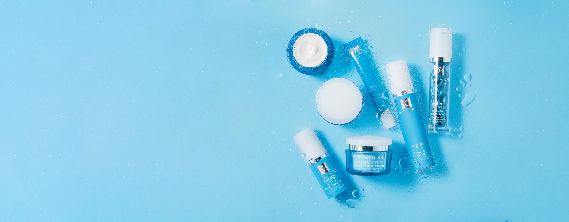 Dr. Grandel Hydro Active Pflegeserie mit verschiedenen blauen Fläschchen, Tuben und Tiegeln, arrangiert auf einem nassen, hellblauen Hintergrund mit Wassertropfen.