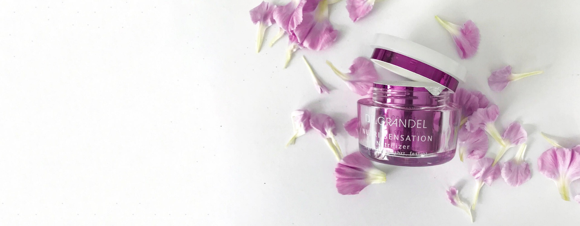 Dr. Grandel Nutri Sensation Nutrilizer Creme in pinkem Tiegel mit geöffnetem Deckel, dekorativ umgeben von violetten Blütenblättern auf weißem Hintergrund.