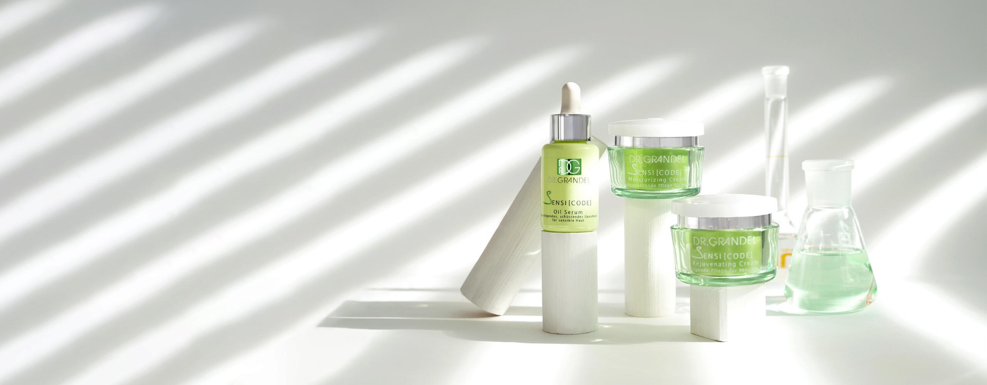 Dr. Grandel Sensicode Pflegeserie mit grünem Oil Serum und zwei Cremes, arrangiert auf weißen Podesten mit Laborgläsern im Hintergrund und Lichtstreifen im Vordergrund.