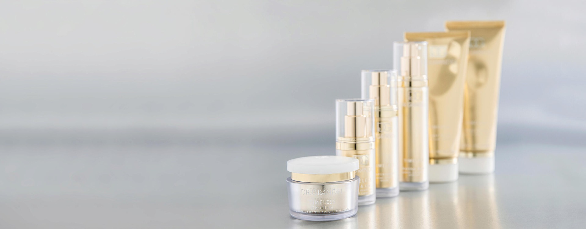 Dr. Grandel Timeless Hautpflegeprodukte in eleganten goldfarbenen Tuben und Flaschen, auf einem hellgrauen Hintergrund ansteigend nach hinten arrangiert.