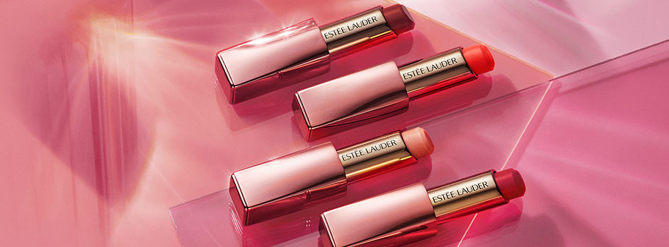 Vier Estée Lauder Lippenstifte in eleganten roségoldenen Hüllen, geöffnet und in verschiedenen Rottönen arrangiert auf glänzendem, pinkfarbenem Untergrund.