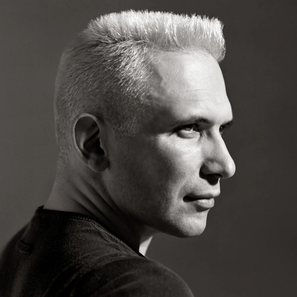 Schwarz-weiß Porträt von Jean Paul Gaultier mit kurzem, hellem Bürstenhaarschnitt in Seitenansicht vor neutralem, dunklen Hintergrund.