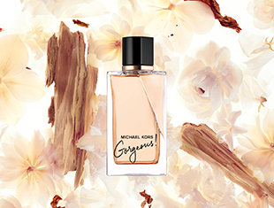 Parfumflakon von 'Michael Kors Gorgeous!' vor einem Hintergrund aus zarten Blüten und Holzstücken. Der eckige Flakon mit schwarzem Deckel enthält einen roséfarbenen Duft. Die Aufschrift auf dem Flakon lautet 'Michael Kors Gorgeous!'.