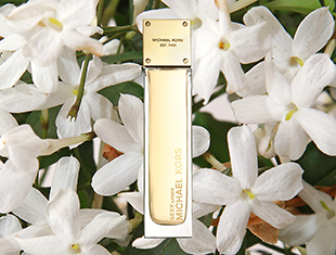 Schmaler, eleganter Parfumflakon von Michael Kors in Gold- und Cremetönen, aufrecht stehend vor einem Hintergrund aus weißen Jasminblüten.