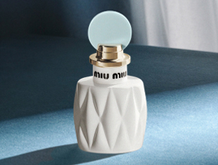 Parfumflakon Miu Miu Fleur de Lait auf blauem Hintergrund. Die weiße, facettierte Flasche trägt eine goldene Fassung mit Miu Miu-Schriftzug und ist mit einem runden, hellblauen Verschluss versehen.