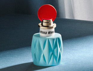 Parfumflakon Miu Miu L’Eau de Muguet auf blauem Hintergrund. Die Flasche ist in einem hellblauen, facettierten Glasdesign gehalten, mit goldener Fassung, weißem Ring mit Miu Miu-Schriftzug und einem auffälligen runden roten Verschluss.