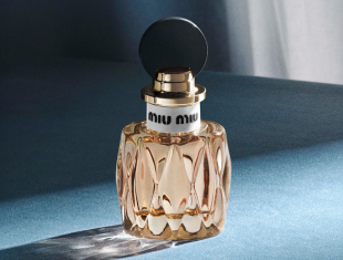 Glasflakon des Parfums Miutine by Miu Miu auf blauem Hintergrund. Die Flasche ist facettenreich geschliffen, enthält einen goldfarbenen Duft, und hat einen runden schwarzen Verschluss mit goldener Fassung.