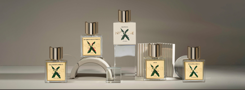 Fünf Parfumflakons der Marke Nishane stehen auf und zwischen transparenten Podesten in einem minimalistischen Setting mit grauem Hintergrund. Die Flakons tragen goldene Etiketten mit grünem 'X' und goldener Kappe, darunter Namen wie 'For Your Flames', 'Hundred Silent Ways' und 'Ani'.
