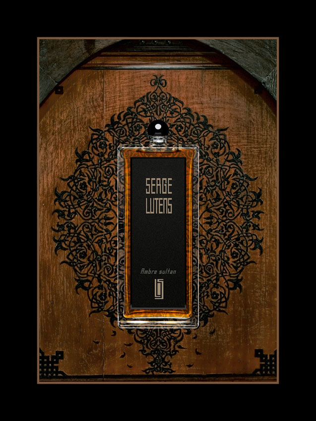 Ein rechteckiger Parfumflakon von Serge Lutens mit schwarzem Etikett und goldener Aufschrift „Ambre Sultan“ steht vor einer dekorativ geschnitzten Holztür mit orientalischem Muster. Die Szene ist kunstvoll inszeniert mit einem schwarzen Rahmen.