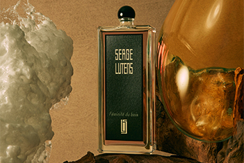 Ein rechteckiger Parfumflakon der Marke Serge Lutens mit schwarzem Etikett und goldener Schrift steht auf einem natürlichen Untergrund. Links davon ist eine große weiße Kristallstruktur, rechts eine bernsteinfarbene Kugel mit organischer Textur zu sehen. Auf dem Etikett steht „Féminité du bois“.