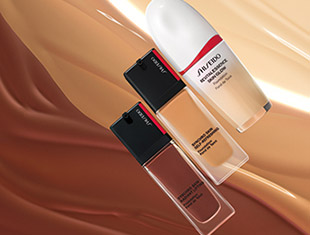 Drei Foundation-Produkte von Shiseido liegen auf einem Hintergrund aus fließenden Make-up-Nuancen in Beige- und Brauntönen. Zwei der Produkte befinden sich in eckigen Glasflakons mit schwarzem Deckel, das dritte in einem weißen, abgerundeten Flakon mit rotem Akzent.