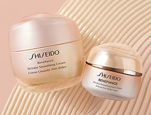 Zwei Tiegel der Shiseido Benefiance Pflegeserie liegen auf einem cremefarbenen, gewellten Untergrund. Links befindet sich die „Wrinkle Smoothing Cream“, rechts die passende Augenpflege „Wrinkle Smoothing Eye Cream“. Die Verpackungen sind in zartem Rosé-Gold mit glänzendem Finish gestaltet.