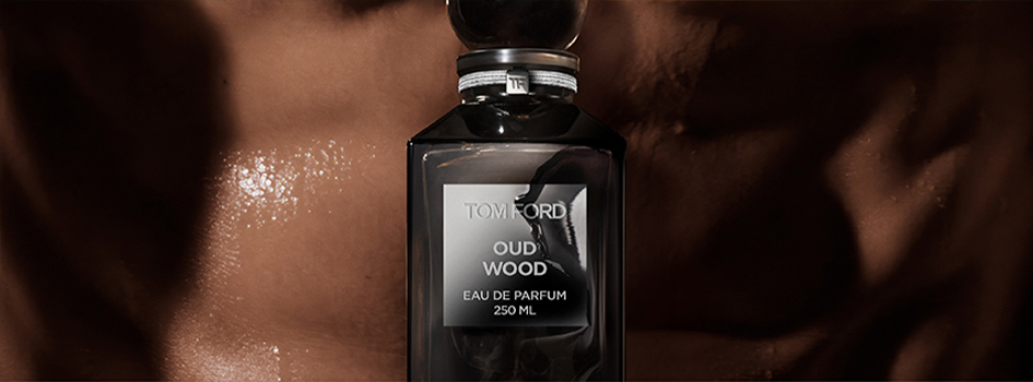 Nahaufnahme eines schwarzen Parfumflakons 'Tom Ford Oud Wood Eau de Parfum 250 ml' vor glänzender, brauner, feucht wirkender Haut. Der Hintergrund erzeugt eine sinnliche, luxuriöse Atmosphäre.